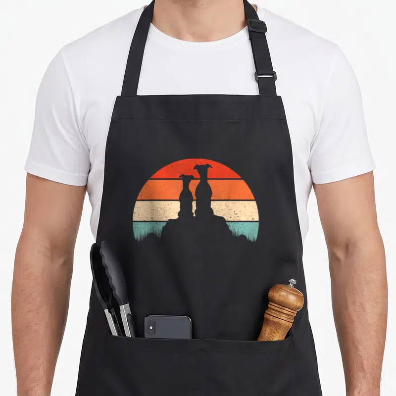 Whippet sunset dog apron - Two black dog silhouettes sitting in grass against a striped retro sun with red, orange — shown on a black cotton canvas apron against a white background