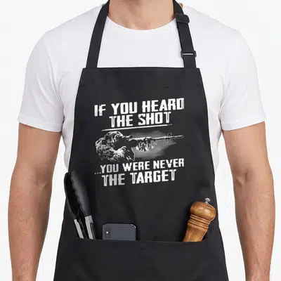 Heard wellness shot apron - Distressed white text reading If You Heard The Shot You Were Never The Target around a grey — shown on a black cotton canvas apron against a white background
