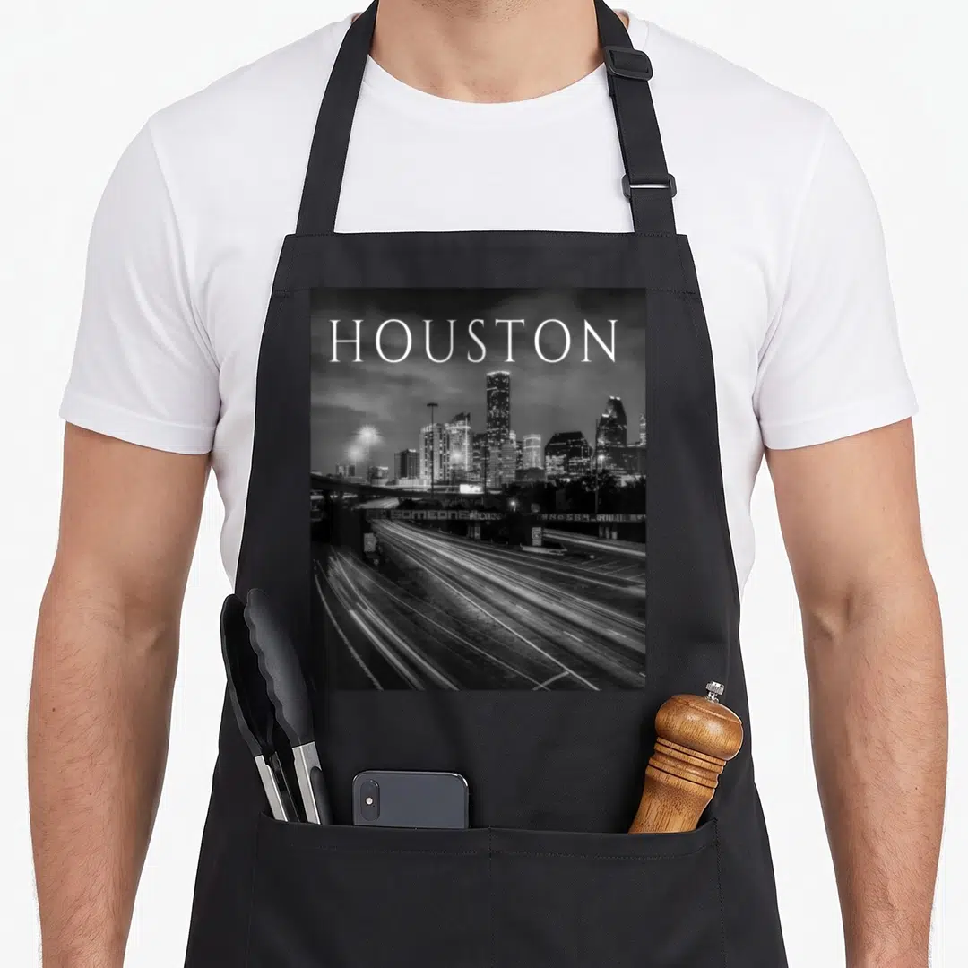 Downtown texas apron - HOUSTON text over black and white skyline and highway light trail photo — shown on a black cotton canvas apron against a white background