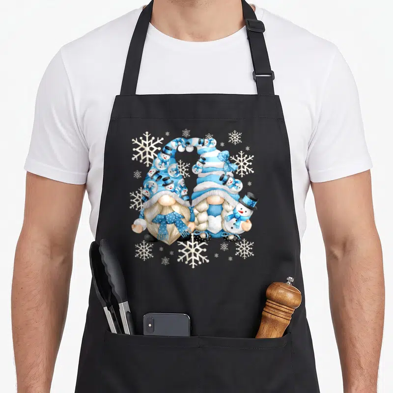 Gnomy snowman apron - Two gnomes with blue striped hats forming a heart surrounded by snowmen and snowflakes — shown on a black cotton canvas apron against a white background