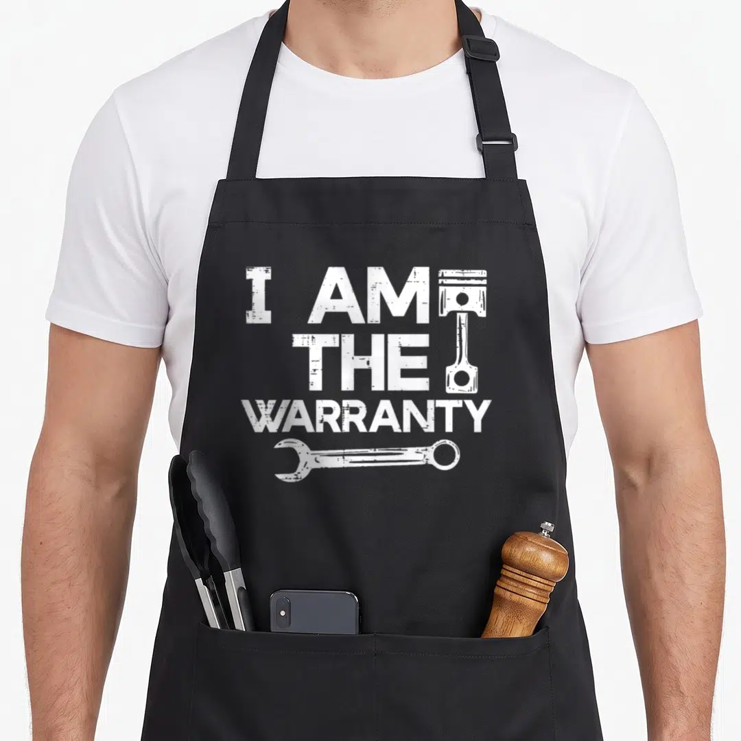 Warranty auto mechanic apron - White distressed text I AM THE WARRANTY with piston and wrench graphics — shown on a black cotton canvas apron against a white background