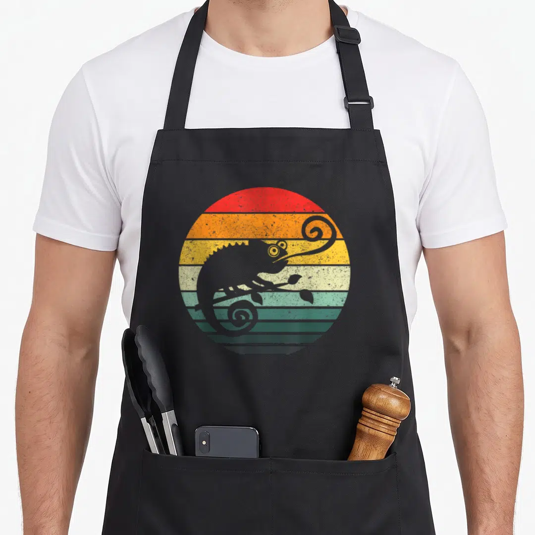 Chameleon animal retro apron - Grey chameleon silhouette perched on branch over circular red orange yellow green grey — shown on a black cotton canvas apron against a white background