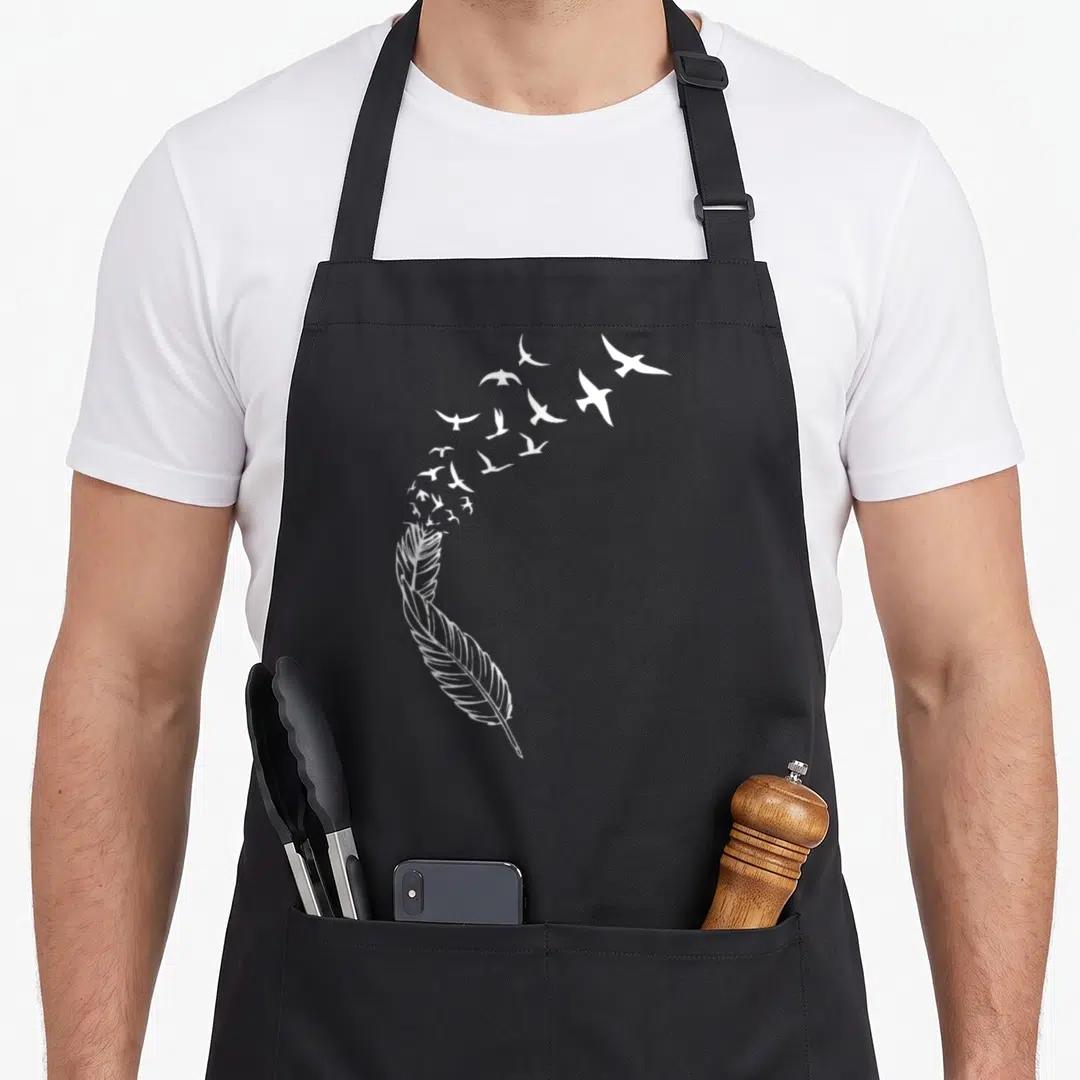 Feather flying apron - White line-art feather with flock of birds flying upward to the right — shown on a black cotton canvas apron against a white background