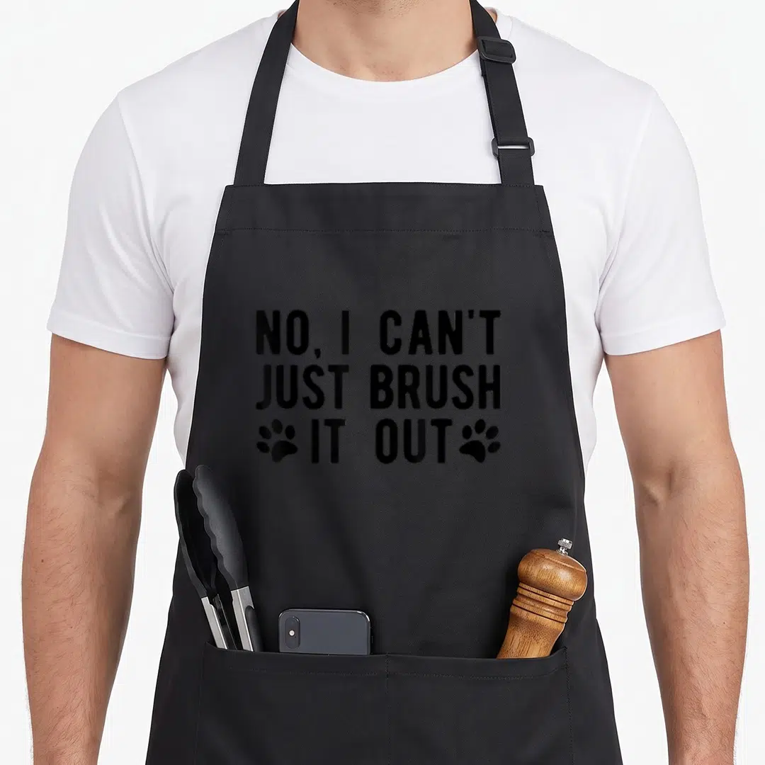 Groomer dog apron - Black text reading No I Can't Just Brush It Out with paw prints on left and right of bottom line — shown on a black cotton canvas apron against a white background