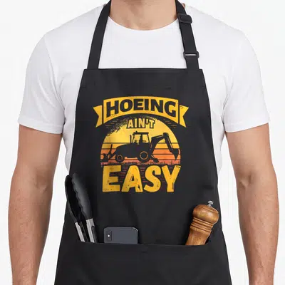 Backhoe easy apron - Retro yellow and orange sun graphic with grey backhoe silhouette and HOEING AIN'T EASY text — shown on a black cotton canvas apron against a white background