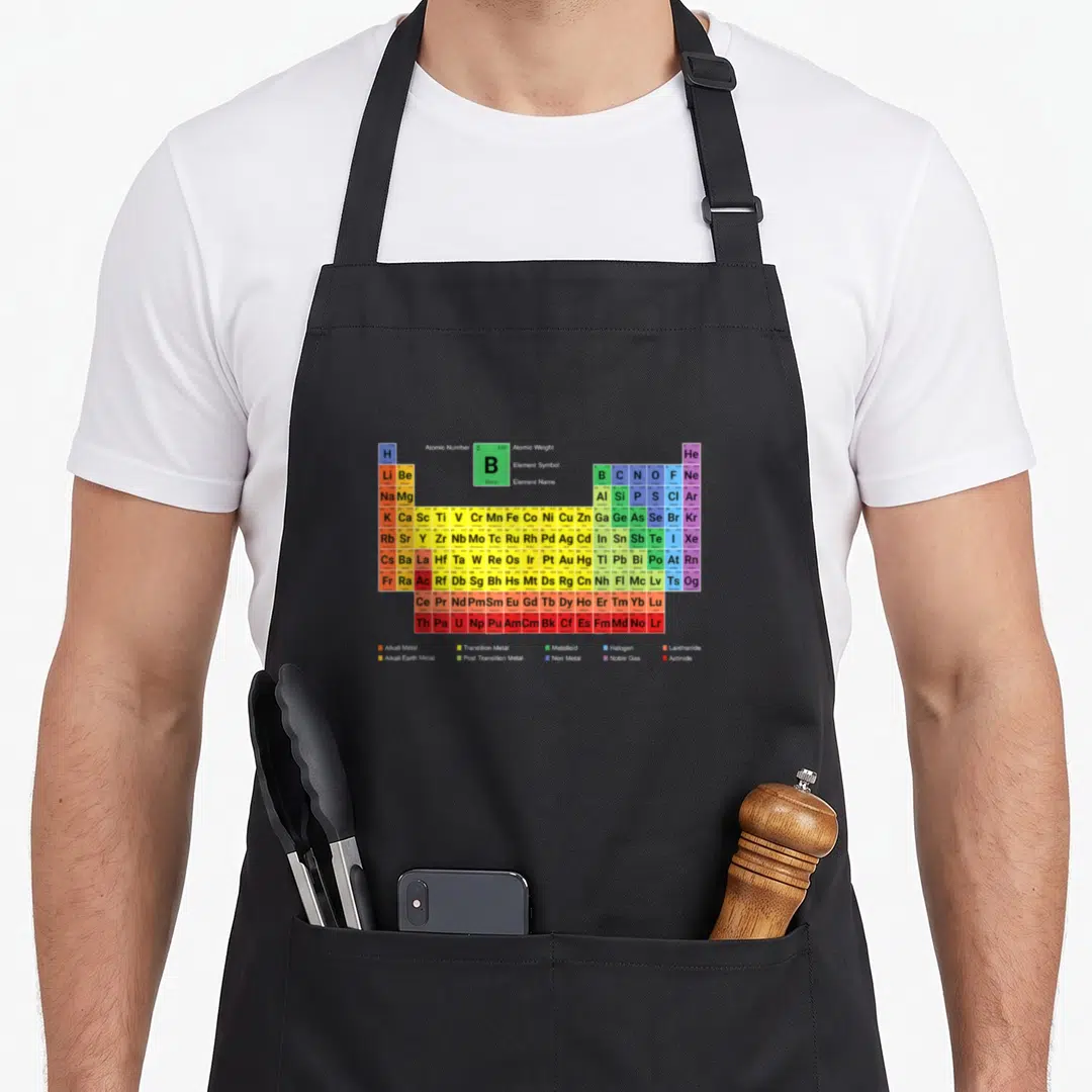 Complete table apron - Periodic table grid with colored boxes, element symbols, and category legend at bottom — shown on a black cotton canvas apron against a white background