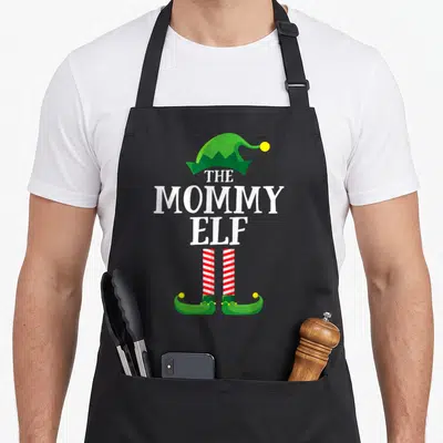 Pyjamas christmas elf apron - Stacked white text THE MOMMY ELF with green hat and red striped legs — shown on a black cotton canvas apron against a white background