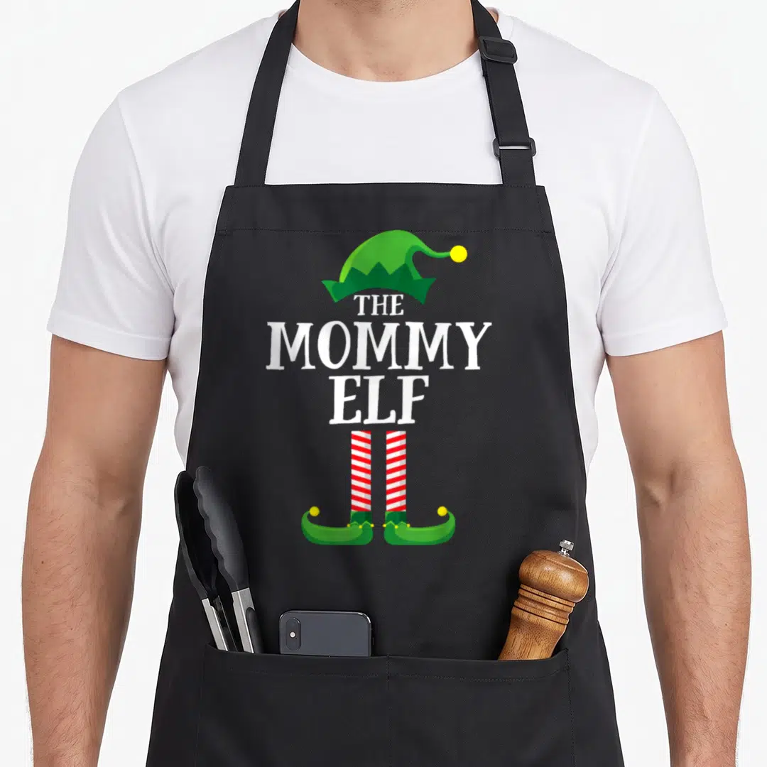 Pyjamas christmas elf apron - Stacked white text THE MOMMY ELF with green hat and red striped legs — shown on a black cotton canvas apron against a white background
