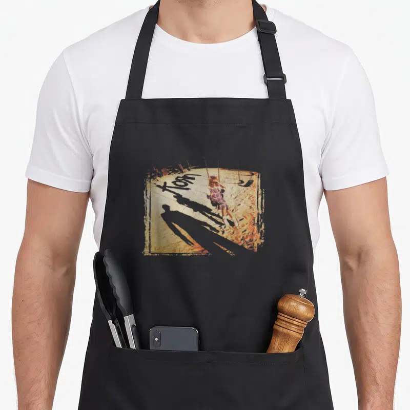 Korn armagnac apron - Distressed illustration of a girl on a swing with a long shadow in red and brown tones — shown on a black cotton canvas apron against a white background