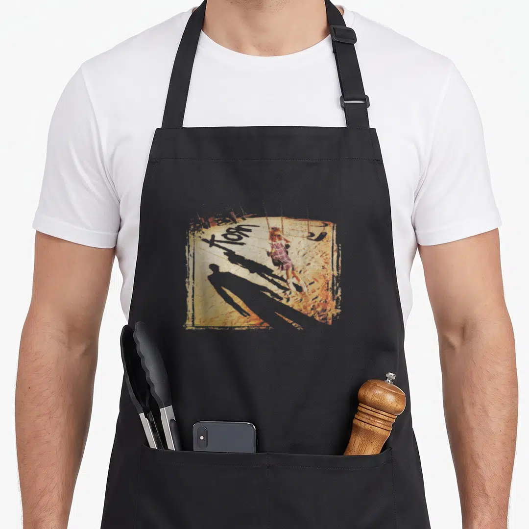 Korn armagnac apron - Distressed illustration of a girl on a swing with a long shadow in red and brown tones — shown on a black cotton canvas apron against a white background