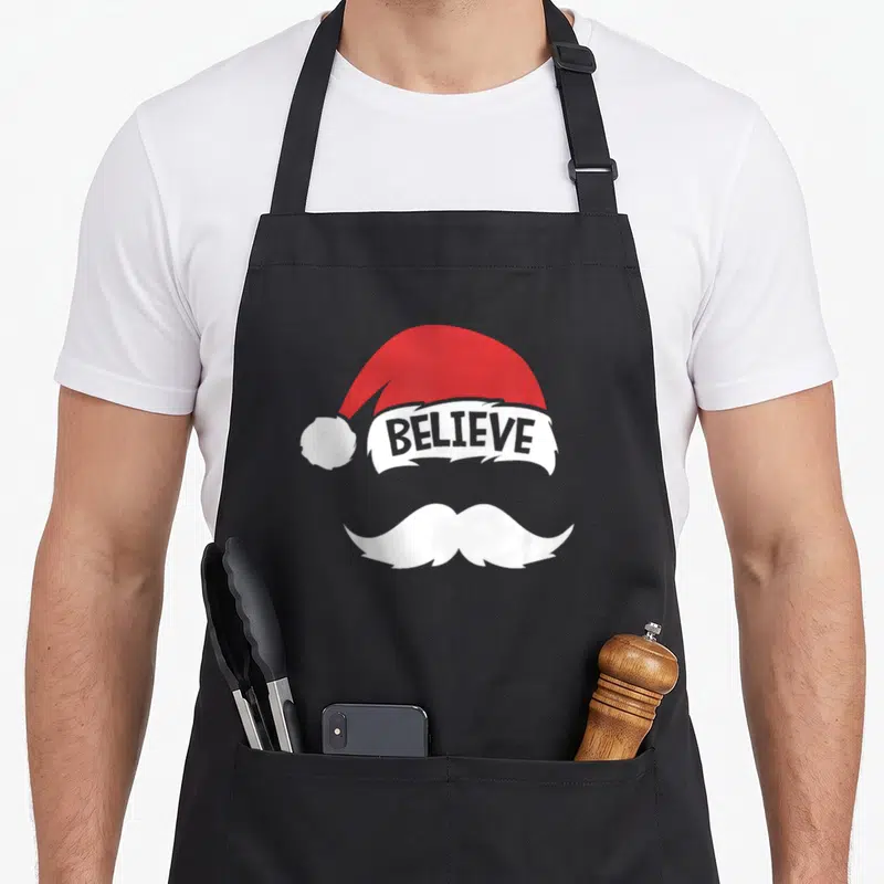 Believe family reunion aprons - Red Santa hat with BELIEVE text on white trim above a white mustache — shown on a black cotton canvas apron against a white background