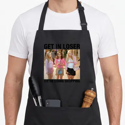 Y2k mean girls apron - Three women photo between GET IN LOSER and WE'RE GOING SHOPPING text — shown on a black cotton canvas apron against a white background