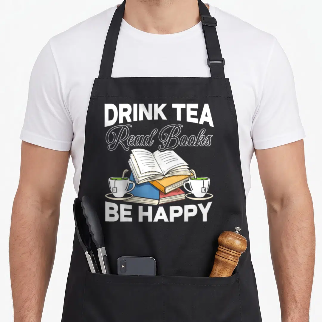 Bibliophile tea apron - Open book on stack flanked by two tea cups with DRINK TEA Read Books BE HAPPY text — shown on a black cotton canvas apron against a white background