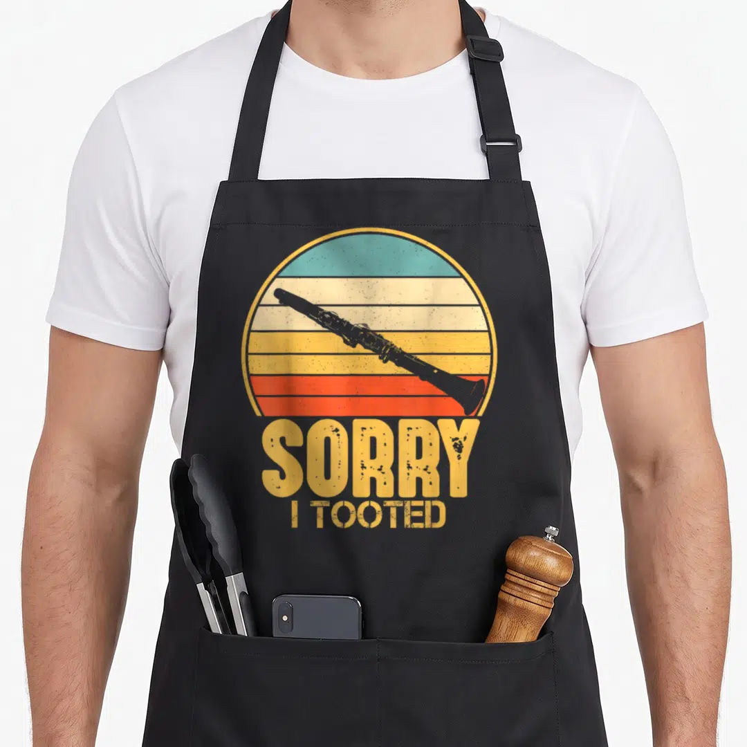 Sorry i love clarinet apron - Grey clarinet on striped yellow orange red teal circle with SORRY I TOOTED text below — shown on a black cotton canvas apron against a white background