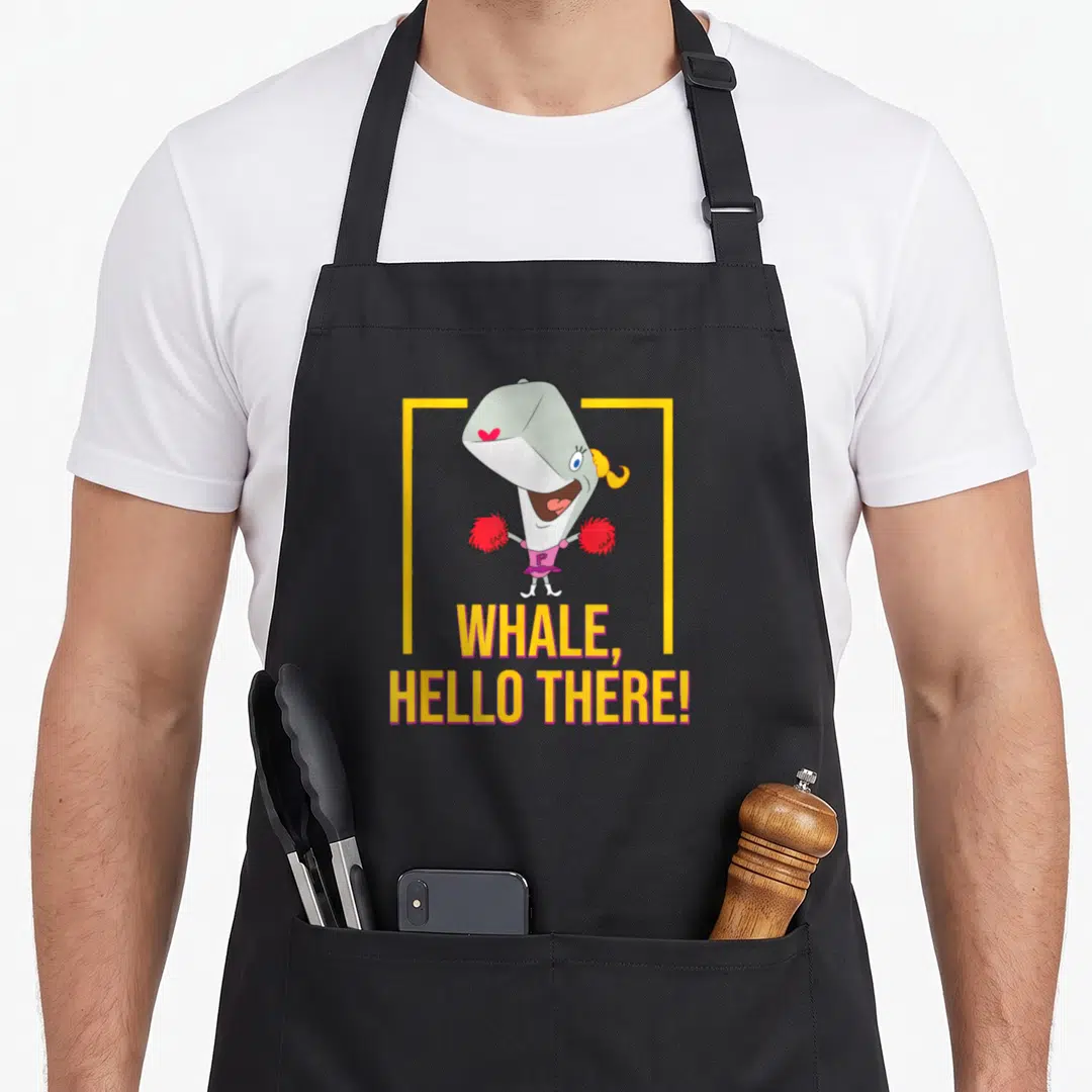 Pearl hello apron - Cartoon grey whale in pink outfit holding red pom-poms with yellow and pink text below — shown on a black cotton canvas apron against a white background