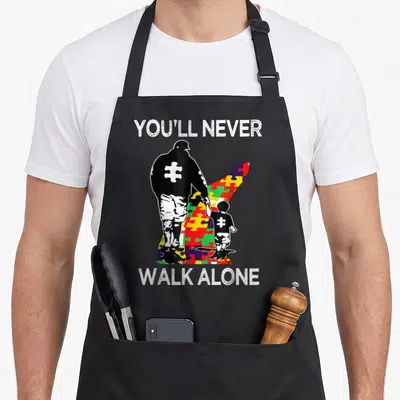 Support autism awareness apron - You'll Never Walk Alone text above and below black man and child silhouettes on a red — shown on a black cotton canvas apron against a white background