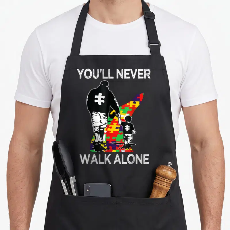 Support autism awareness apron - You'll Never Walk Alone text above and below black man and child silhouettes on a red — shown on a black cotton canvas apron against a white background