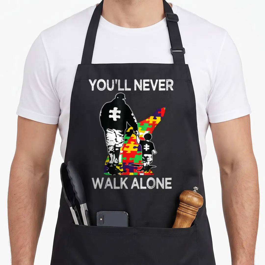 Support autism awareness apron - You'll Never Walk Alone text above and below black man and child silhouettes on a red — shown on a black cotton canvas apron against a white background