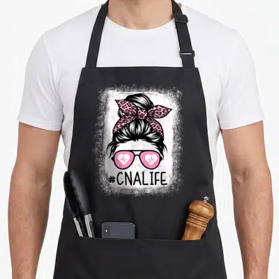 Cna messy aprons - Cartoon woman head with pink leopard bow, heart EKG sunglasses, and #CNALIFE text — shown on a black cotton canvas apron against a white background