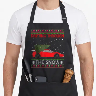 Drifting ugly christmas sweater apron - Red car carrying Christmas tree with drifting through the snow text — shown on a black cotton canvas apron against a white background