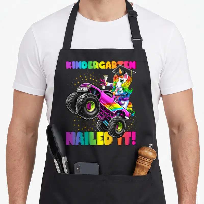 Nailed unicorn aprons - Rainbow unicorn in graduation cap driving pink monster truck with KINDERGARTEN NAILED IT text — shown on a black cotton canvas apron against a white background