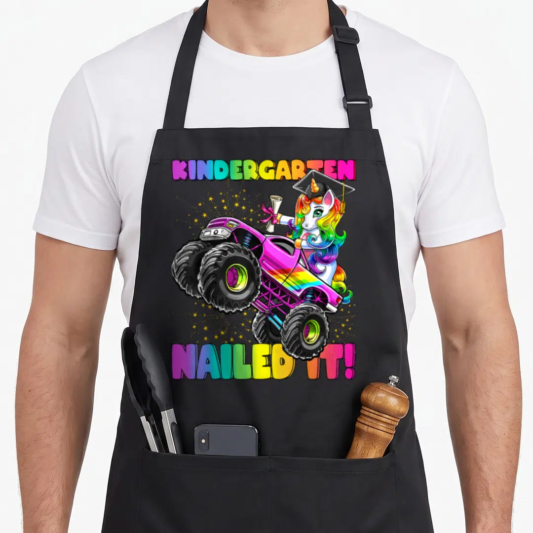 Nailed unicorn aprons - Rainbow unicorn in graduation cap driving pink monster truck with KINDERGARTEN NAILED IT text — shown on a black cotton canvas apron against a white background