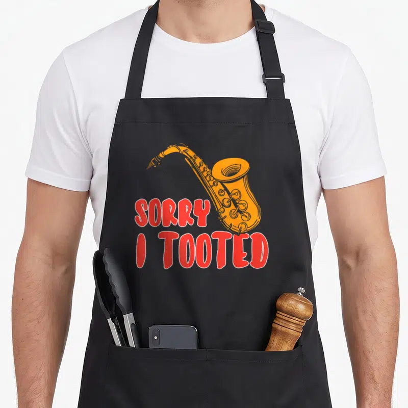 Sorry saxophone apron - Orange cartoon saxophone overlapping red SORRY I TOOTED text with white outlines — shown on a black cotton canvas apron against a white background