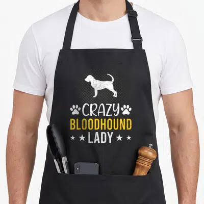 Bloodhound pup apron - White bloodhound silhouette on grey halftone circle with stacked white and yellow text reading — shown on a black cotton canvas apron against a white background