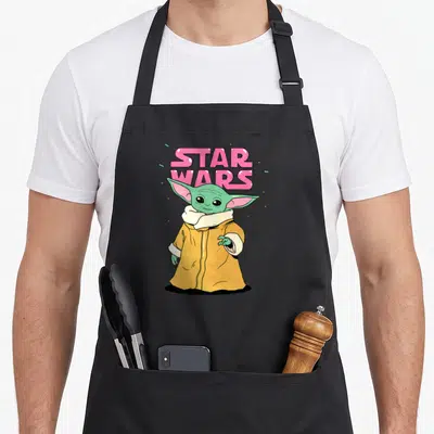 Letter bubble apron - Pink STAR WARS text above cartoon Grogu in green robe and tan coat — shown on a black cotton canvas apron against a white background