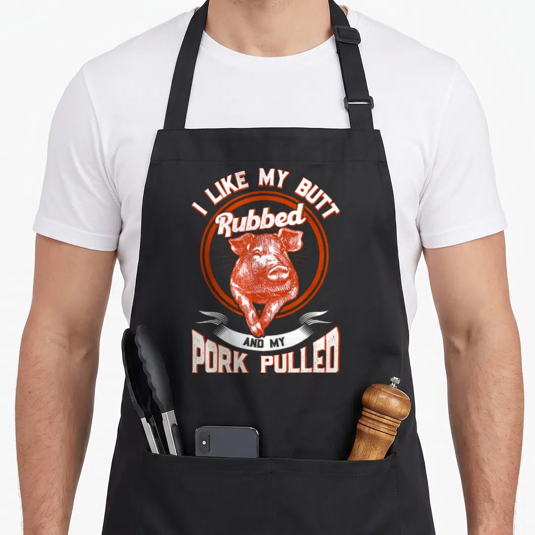 Butt funny barbecue apron - Retro pig illustration with text I Like My Butt Rubbed And My Pork Pulled in orange — shown on a black cotton canvas apron against a white background