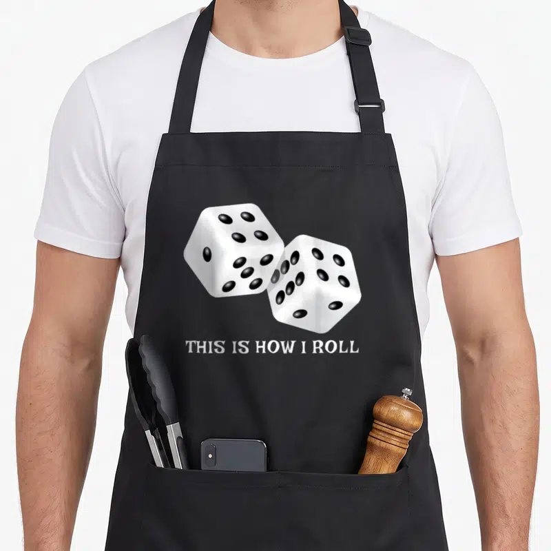 Board roll game aprons - Two white dice with black dots above white text reading THIS IS HOW I ROLL — shown on a black cotton canvas apron against a white background