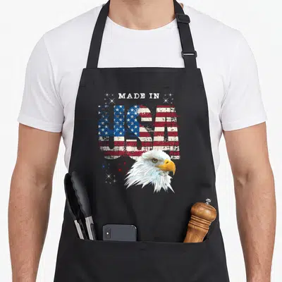 Patriotic bald eagle american flag apron - MADE IN USA text filled with flag pattern and bald eagle head in red white — shown on a black cotton canvas apron against a white background