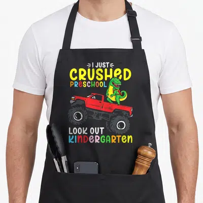 Crushed preschool apron - Green dinosaur in red monster truck with I Just Crushed Preschool Look Out Kindergarten text — shown on a black cotton canvas apron against a white background