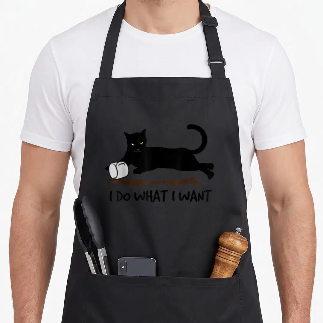 General kitty apron - Black cat knocking over white mug with brown spill and text I DO WHAT I WANT — shown on a black cotton canvas apron against a white background