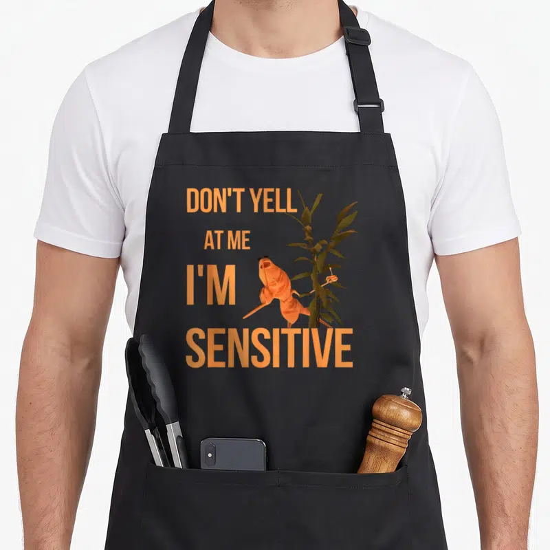 Marcus meme apron - Orange creature with sunglasses and plant beside text Don't Yell At Me I'm Sensitive — shown on a black cotton canvas apron against a white background