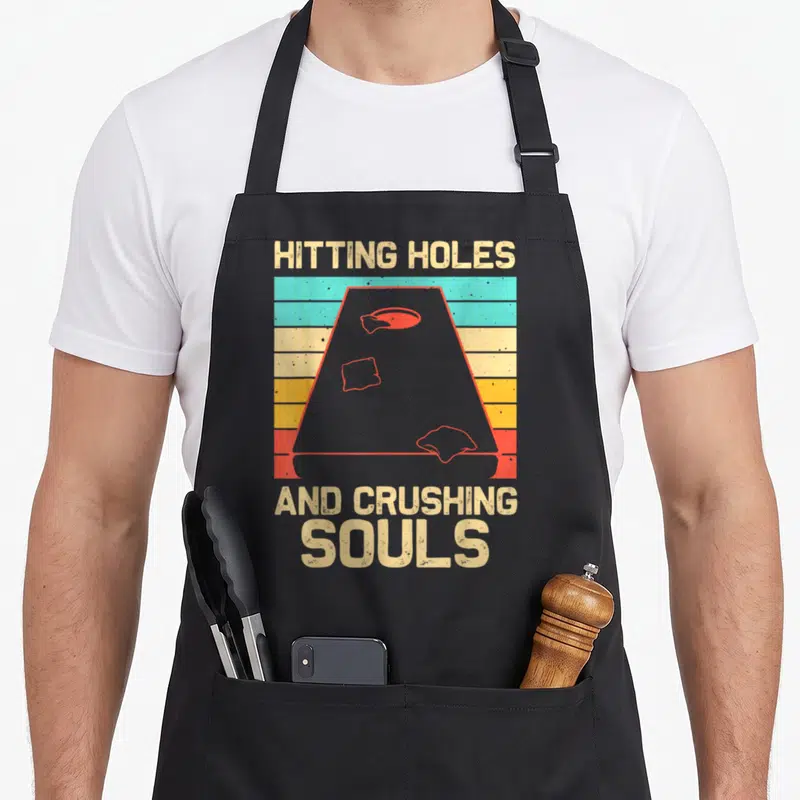 Player hole apron - HITTING HOLES AND CRUSHING SOULS text above grey trapezoid with red outlines over colored stripes — shown on a black cotton canvas apron against a white background