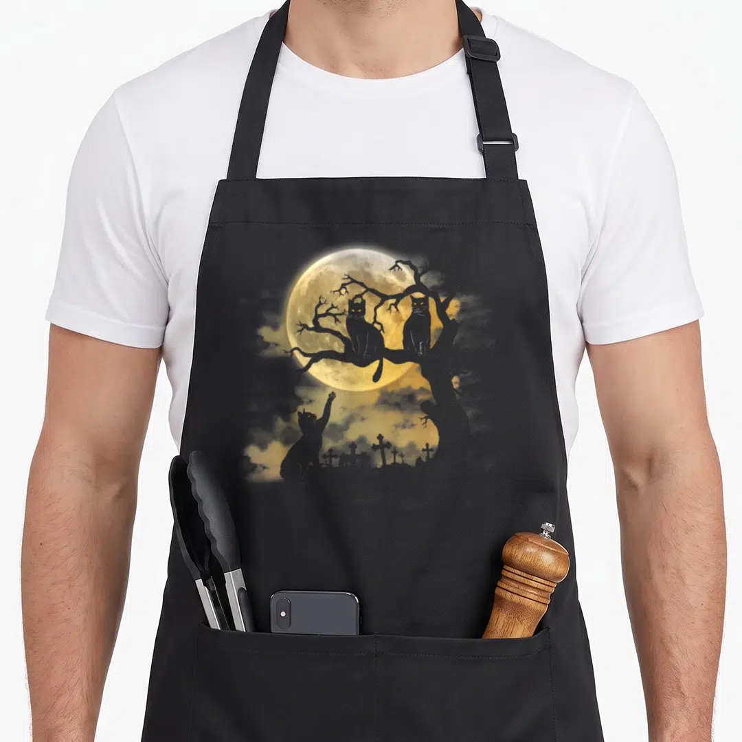 Cat spooky apron - Three grey cats on a branch and graveyard under a yellow full moon — shown on a black cotton canvas apron against a white background