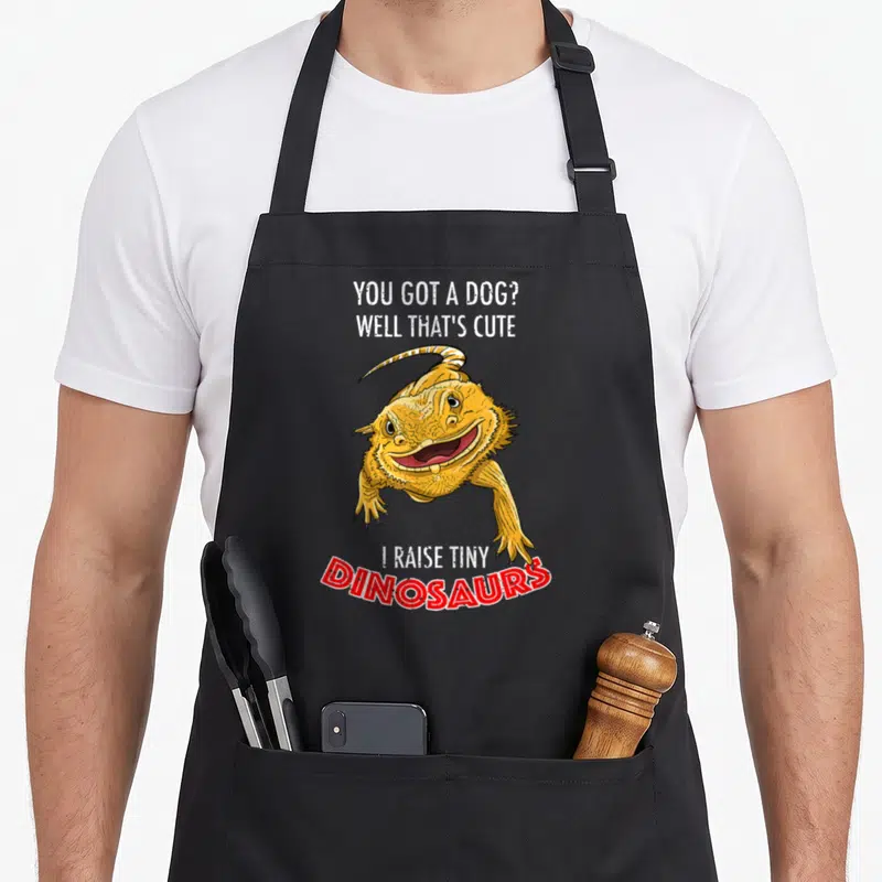 Dinosaur tiny chef apron - Yellow cartoon bearded dragon surrounded by text You Got A Dog Well That's Cute I Raise Tiny — shown on a black cotton canvas apron against a white background