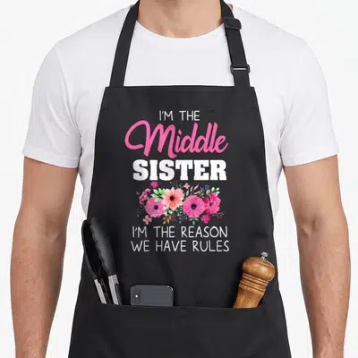 Sister middle squad apron - I'M THE Middle SISTER text with pink and green flower cluster — shown on a black cotton canvas apron against a white background