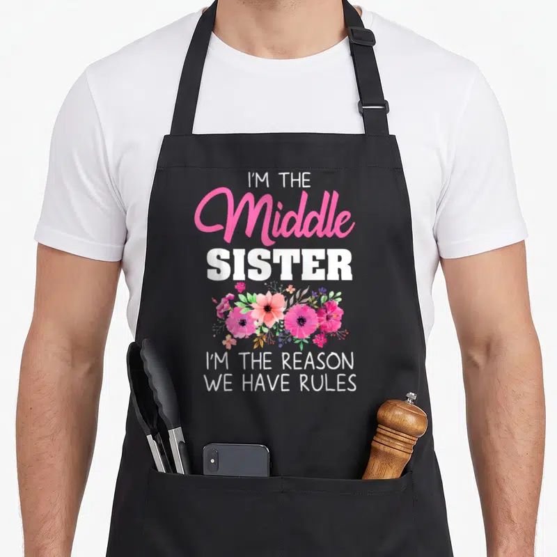 Sister middle squad apron - I'M THE Middle SISTER text with pink and green flower cluster — shown on a black cotton canvas apron against a white background