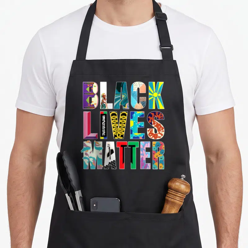 Live celebrate diversity apron - Stacked BLACK LIVES MATTER text letters filled with colorful unique illustrations — shown on a black cotton canvas apron against a white background