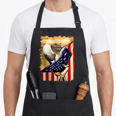 Flag fight rooster apron - Realistic rooster and hand holding draped red white blue flag in front of barn — shown on a black cotton canvas apron against a white background