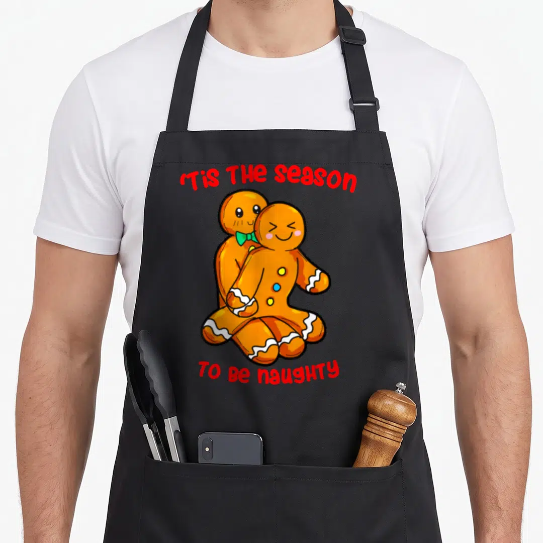 Tis gingerbread apron - Two hugging gingerbread men with red text Tis The season TO BE naughty — shown on a black cotton canvas apron against a white background