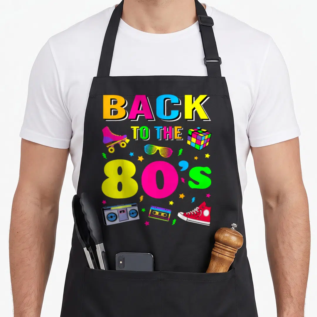 Eighty costume 80s apron - Stacked BACK TO THE 80'S text with boombox, skate, cube, glasses, tape, shoe, and stars — shown on a black cotton canvas apron against a white background