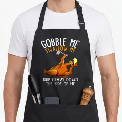 Swallow thanksgiving apron - Cartoon roasted turkey with text GOBBLE ME SWALLOW ME DRIP GRAVY DOWN THE SIDE OF ME — shown on a black cotton canvas apron against a white background