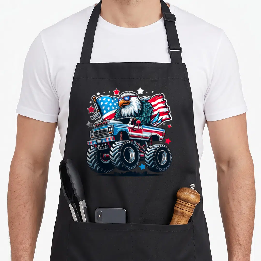 Flag eagle 4th of july apron - Cartoon bald eagle in monster truck with USA flag and stars in red white blue — shown on a black cotton canvas apron against a white background