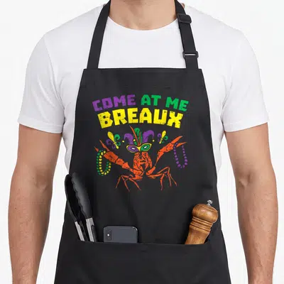 Come mardi gras aprons - Come At Me Breaux text over jester crawfish with beads and fleur-de-lis — shown on a black cotton canvas apron against a white background