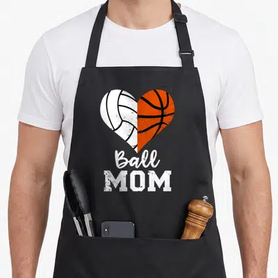 Ball basketball mom apron - Split heart with white volleyball and orange basketball above Ball MOM text — shown on a black cotton canvas apron against a white background