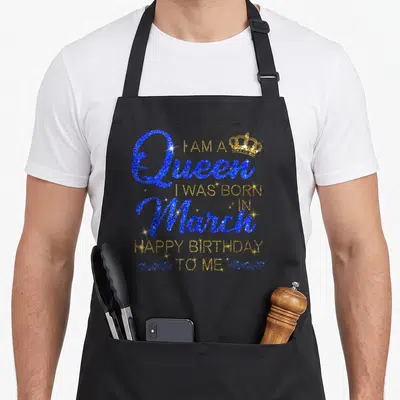 Funny march baking queen apron - Blue and gold typography reading I am a Queen born in March Happy Birthday To Me — shown on a black cotton canvas apron against a white background