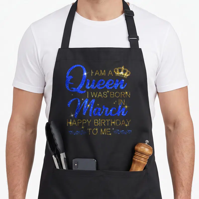 Funny march baking queen apron - Blue and gold typography reading I am a Queen born in March Happy Birthday To Me — shown on a black cotton canvas apron against a white background